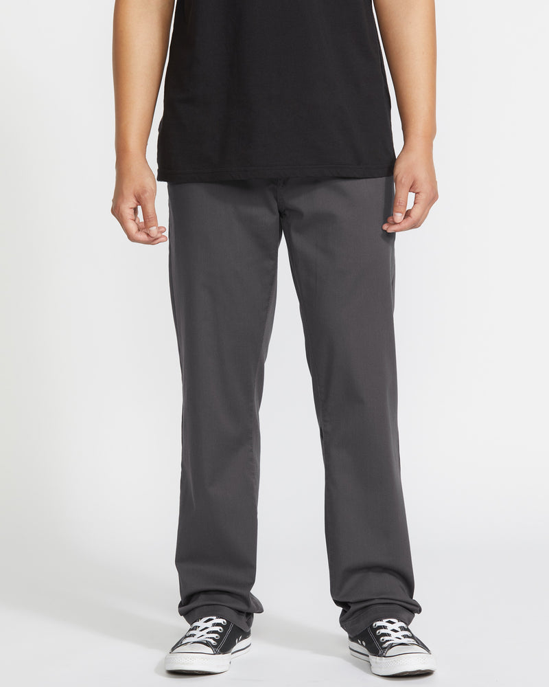 A person wears a black T-shirt, Volcom Men's Frickin Modern Stretch Pants in dark gray, and black-and-white sneakers, standing against a plain white background. The image is cropped at the shoulders.