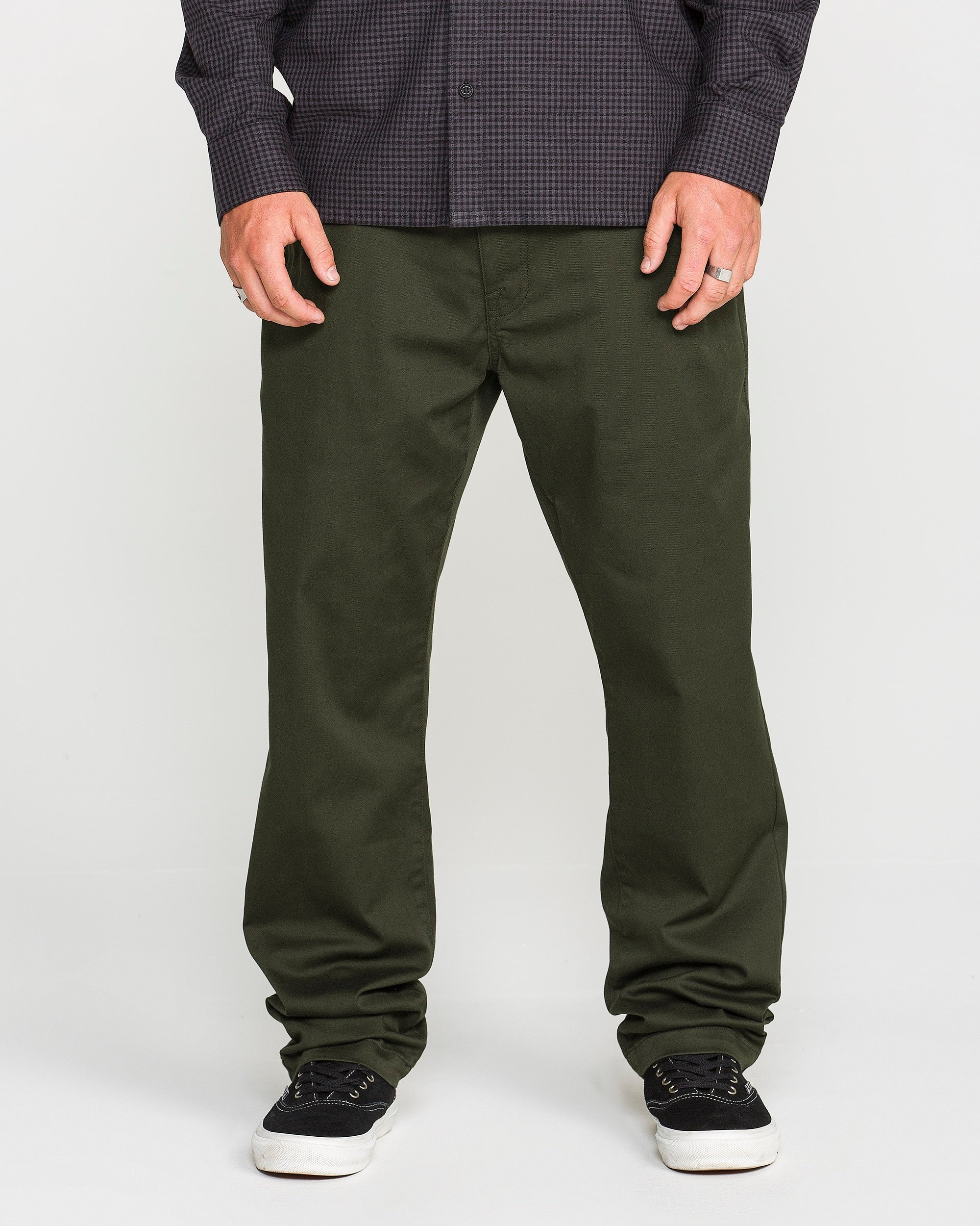 A person wearing Volcom's Men's Frickin Modern Stretch Pants in dark green, crafted with REPREVE recycled fibers, paired with a dark checkered Volcom shirt and black sneakers with white soles, standing against a plain light background.