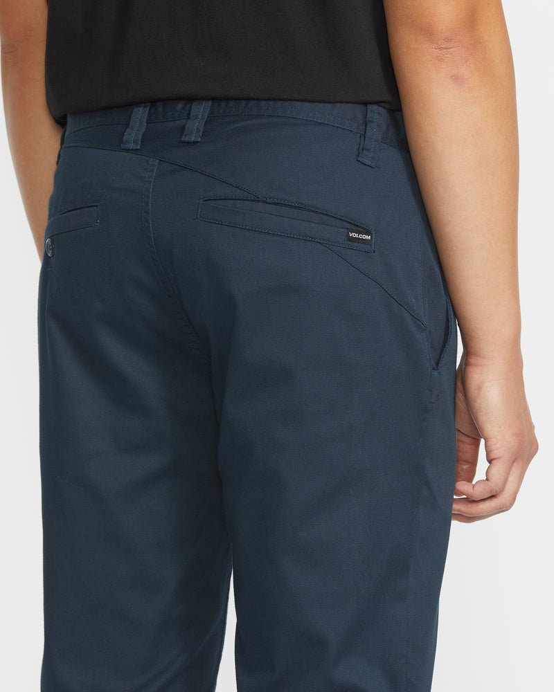 A person is shown from the waist down, viewed from behind, wearing Volcom Men's Frickin Modern Stretch Pants in dark blue and a black shirt. The pants have five-pocket styling and a Volcom label on one back pocket.