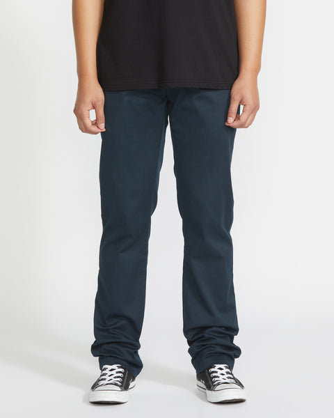 A person wearing a black t-shirt, Volcom Men's Frickin Modern Stretch Pants in dark blue with five-pocket styling, and black-and-white sneakers stands against a plain white background, visible from shoulders to feet. Hands are relaxed by their sides.