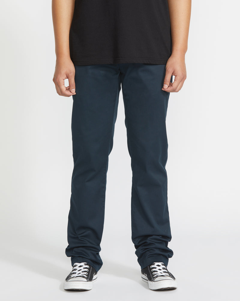A person wearing a black t-shirt, Volcom Men's Frickin Modern Stretch Pants in dark blue with five-pocket styling, and black-and-white sneakers stands against a plain white background, visible from shoulders to feet. Hands are relaxed by their sides.