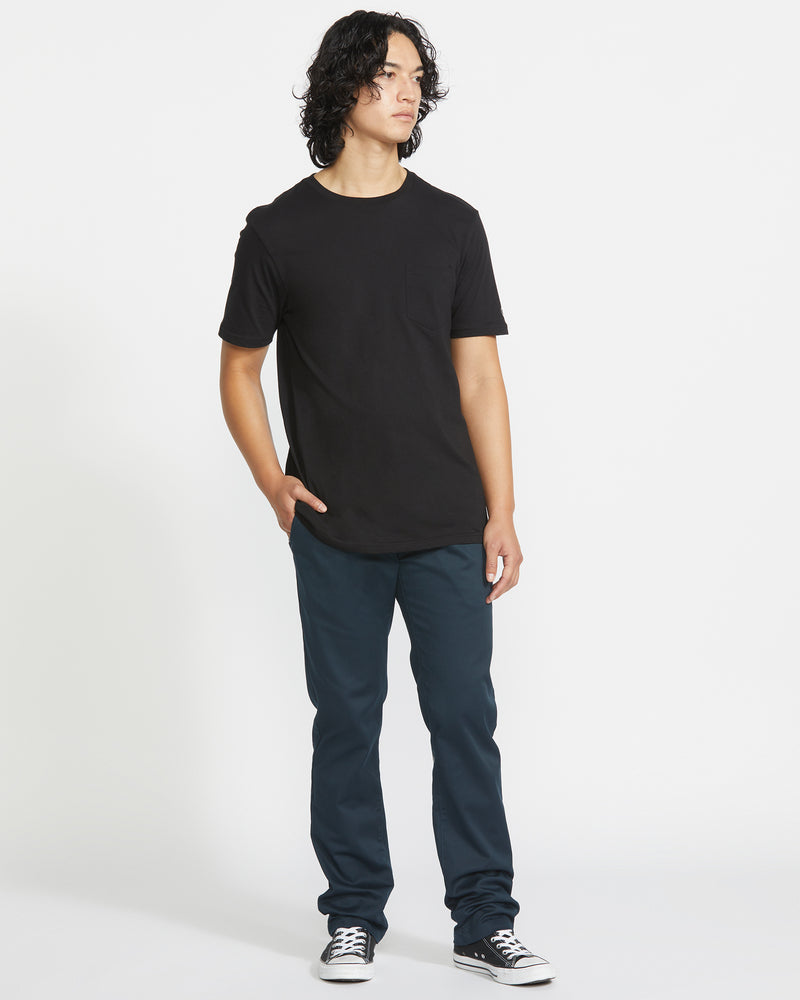 A person with medium-length curly hair stands against a white background, wearing a black T-shirt, Volcom Men's Frickin Modern Stretch Pants in dark blue, and black sneakers with white soles; hands in pockets, looking slightly to the side.