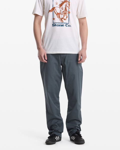 A person stands against a plain white background wearing a white graphic horse T-shirt, Volcom Men's Frickin Modern Stretch Pants in blue-gray, and black Adidas sneakers.
