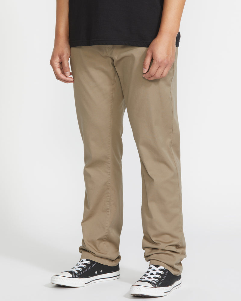 A person wearing Volcom Men’s Frickin Modern Stretch Pants in tan, paired with a black shirt and black low-top sneakers, stands against a plain white background. Only their lower body is visible; the pants feature recycled polyester for an eco-friendly touch.