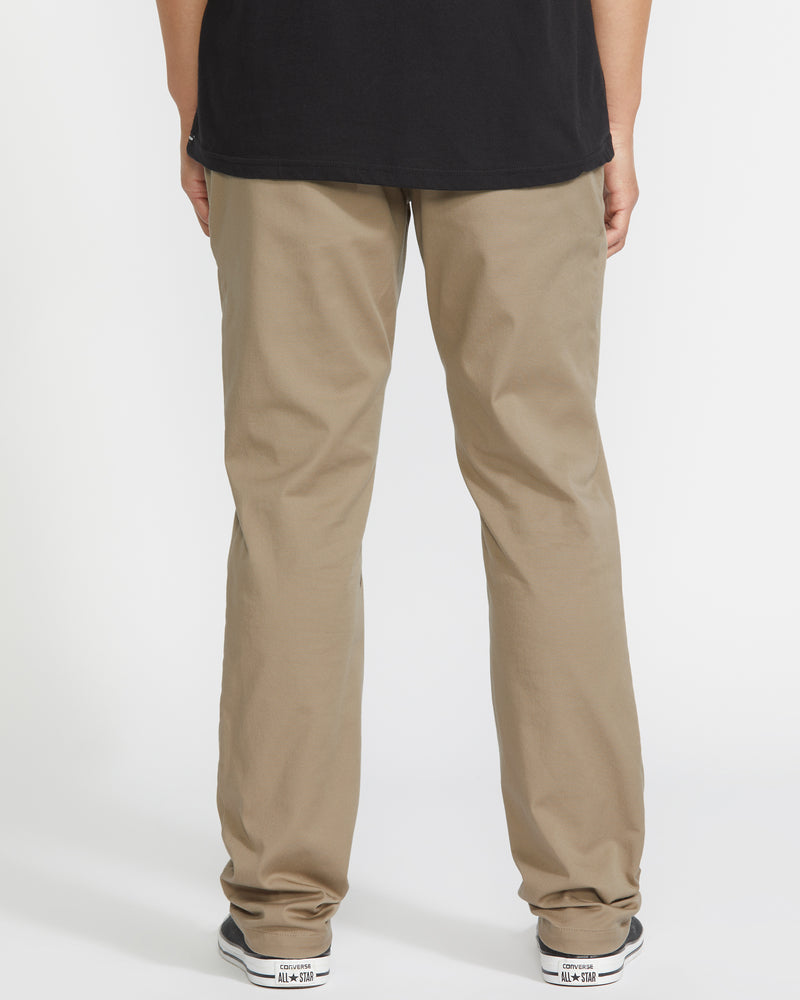 A person wearing Volcom Men’s Frickin Modern Stretch Pants, a black shirt, and white sneakers stands facing away from the camera against a white background.