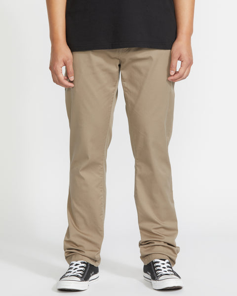 A person wearing Volcom Men’s Frickin Modern Stretch Pants in khaki, paired with a black T-shirt and black-and-white sneakers, stands against a plain white background. Only the lower half of their body is visible. These eco-friendly pants feature recycled polyester for comfortable wear.