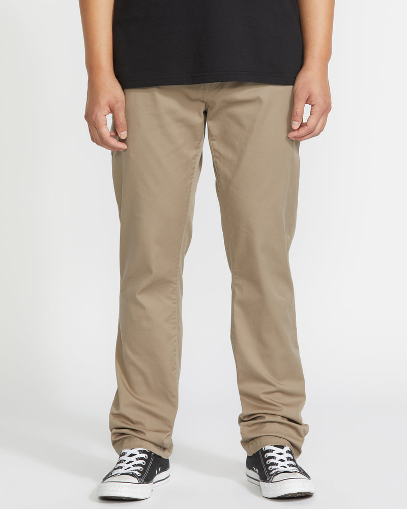 A person wearing Volcom Men’s Frickin Modern Stretch Pants in khaki, paired with a black T-shirt and black-and-white sneakers, stands against a plain white background. Only the lower half of their body is visible. These eco-friendly pants feature recycled polyester for comfortable wear.