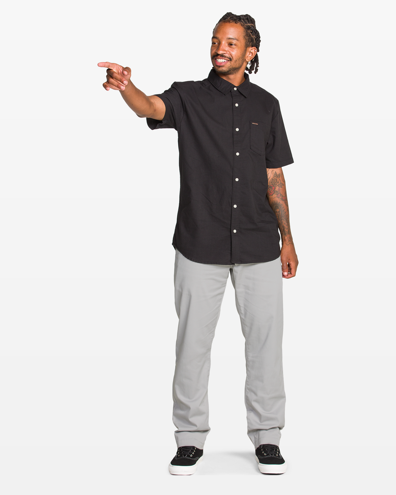 A man with braided hair, wearing a Volcom black short-sleeve button-up shirt and Volcom Men's Frickin Modern Stretch Pants in light gray, stands smiling and pointing to his left against a plain white background.