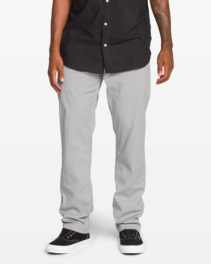 A person wearing Volcom Men's Frickin Modern Stretch Pants, a black button-up shirt, and black sneakers with white soles stands against a plain white background. Only the lower body is visible.
