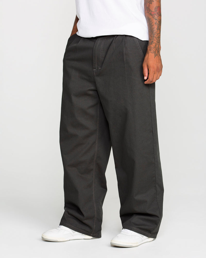 A person is seen from the waist down wearing Volcom Men's Baggos Pleat Pants in a loose, baggy fit styled like chinos. The outfit is paired with white sneakers and a white T-shirt, showing tattoos on their right forearm. The plain, light-colored background reflects a relaxed Volcom aesthetic.