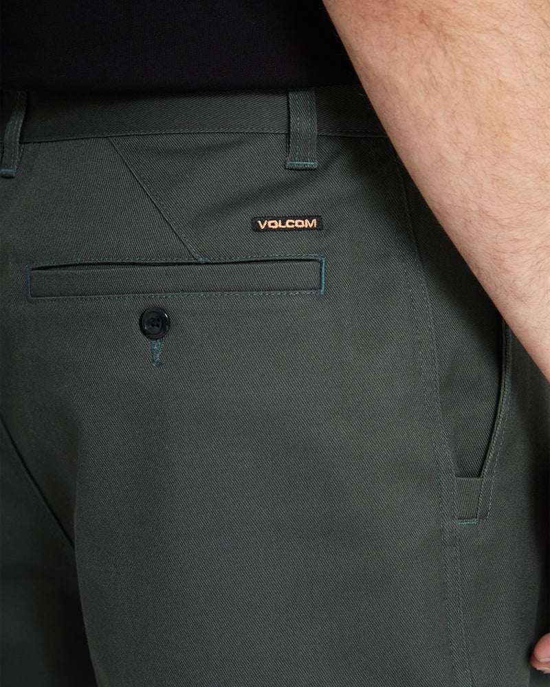 Close-up of a person wearing Volcom Men's Billow Pants in dark green, highlighting the back pocket with a button, a belt loop, and the Volcom logo tag. Part of their arm and black shirt are also visible.