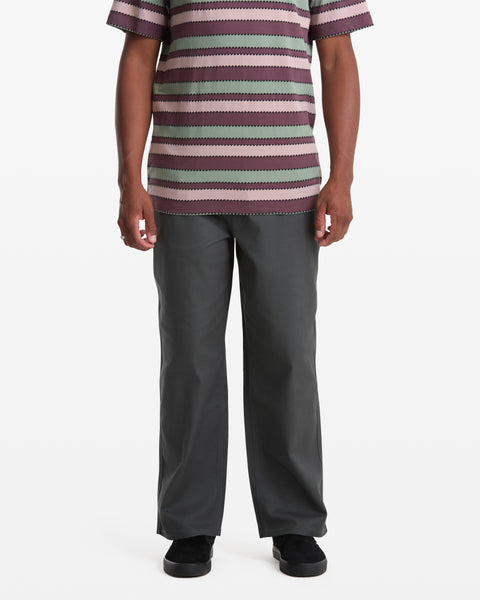 A man stands facing forward in Volcom Men’s Billow Pants, paired with a short-sleeve, striped shirt and black shoes against a plain white background. The image is cropped at the shoulders.