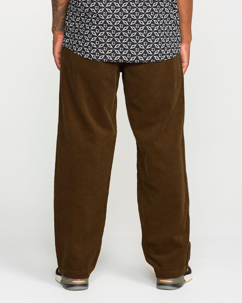 A person stands facing away against a plain light background, wearing Volcom's Men's Frickin Loose Tapered Cord pants in a rich Desert Palm shade. The pants, crafted from 100% cotton corduroy for comfort and style, are paired with a black and white patterned shirt and black shoes featuring white soles.