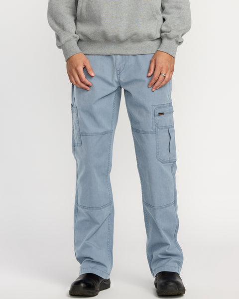 A person stands against a plain white background wearing Volcom Men's Gage Work Pants in light blue, featuring multiple pockets and visible stitching, paired with a gray sweatshirt and black shoes.