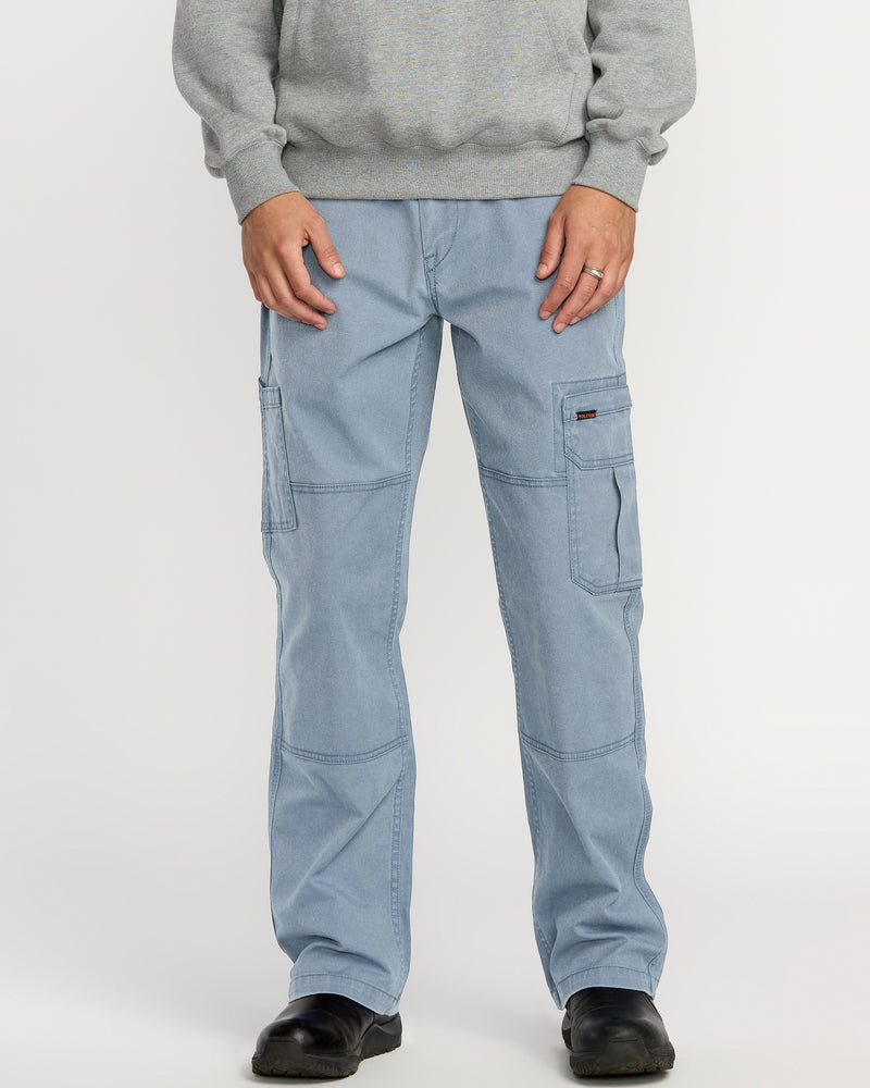 A person stands against a plain white background wearing Volcom Men's Gage Work Pants in light blue, featuring multiple pockets and visible stitching, paired with a gray sweatshirt and black shoes.