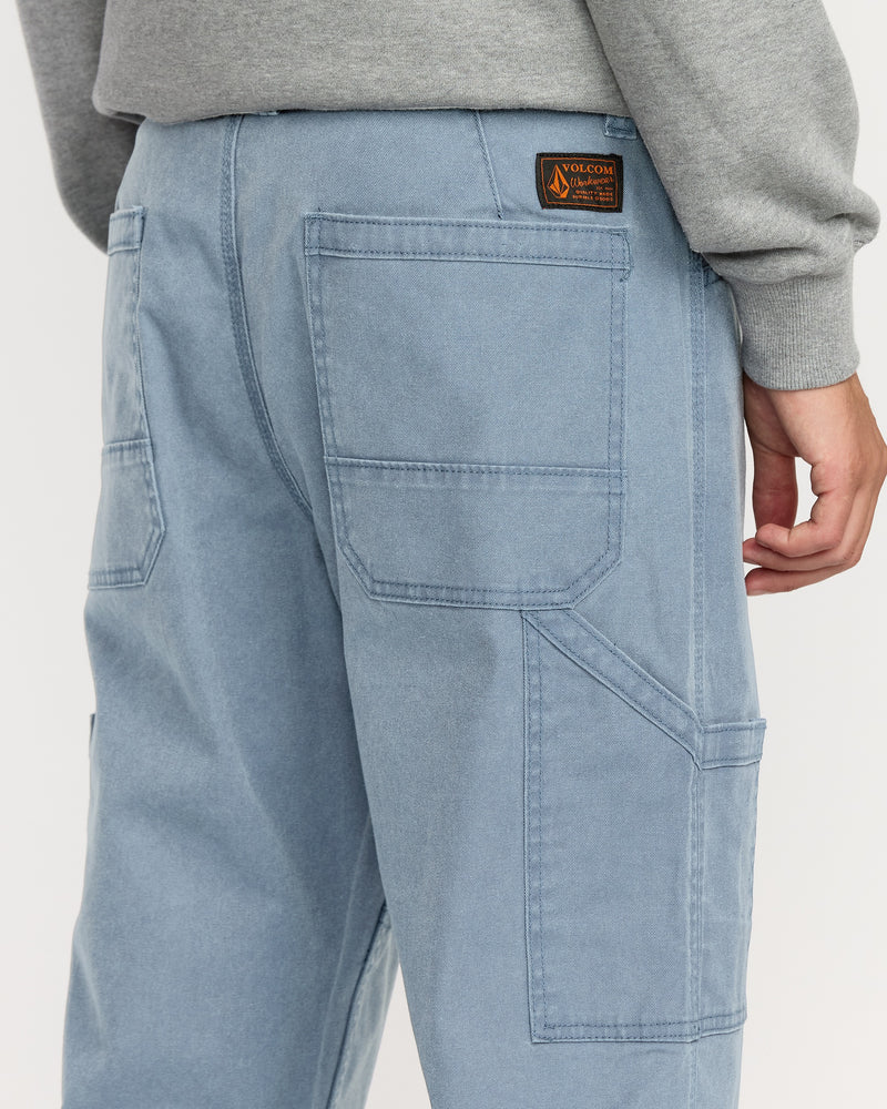 A person wearing Volcom Men's Gage Work Pants in light blue denim, featuring large back pockets and a branded patch near the waistband; a gray sweatshirt sleeve is partially visible.