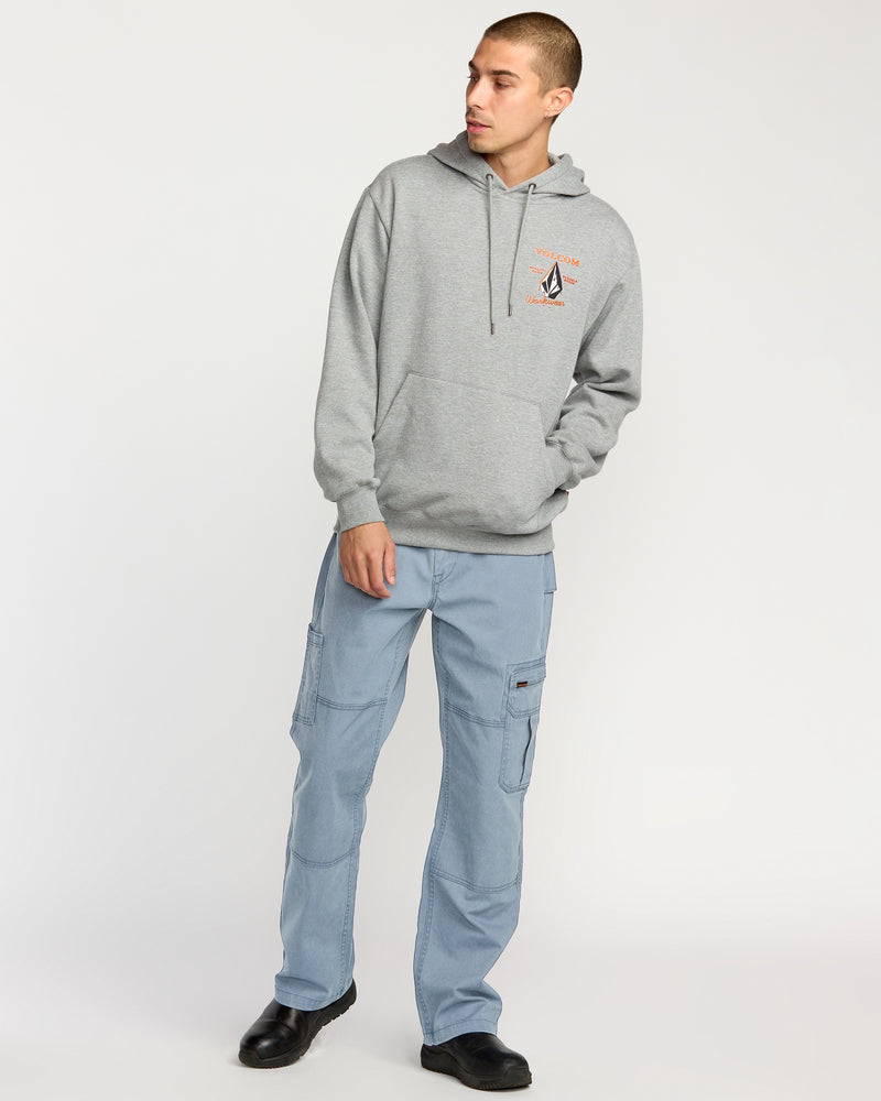 A person stands against a plain background wearing light blue Volcom Men's Gage Work Pants, a gray hooded sweatshirt with a small chest graphic, and black shoes, looking to the side with one hand in their pocket.