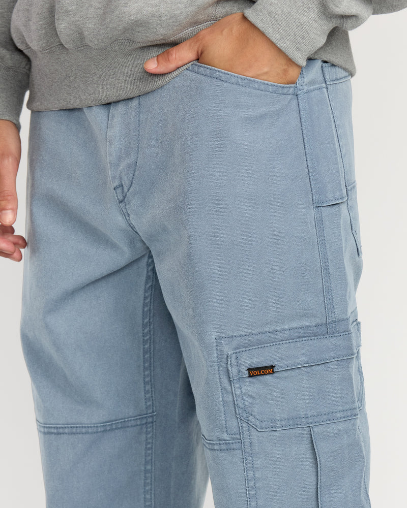 A person wears Volcom Men's Gage Work Pants in light blue, featuring visible stitching and a small logo tag on the side pocket, paired with a gray sweatshirt and one hand in their pocket.