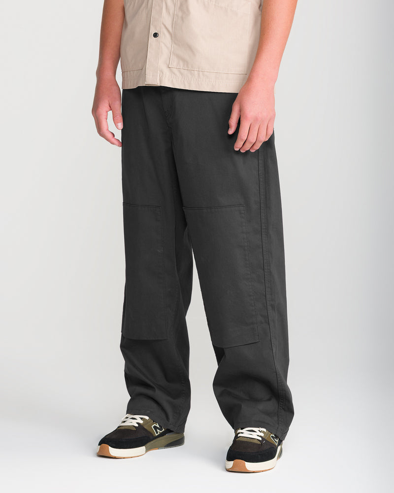 A person wearing the Volcom Men’s Stoneworks Utility Work Pant, featuring reinforced front-leg patches, stands against a plain light background. The outfit is paired with black sneakers with white laces and beige soles, and a beige short-sleeve shirt.
