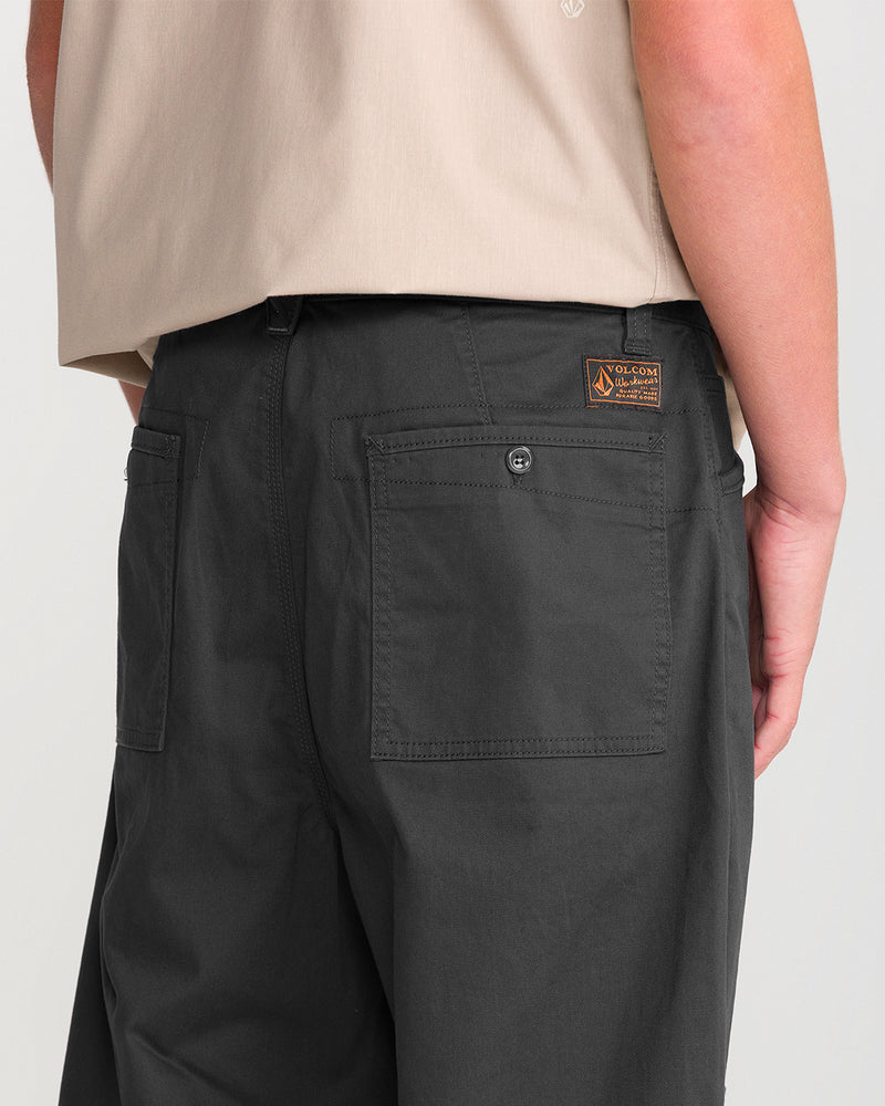 A person wearing Volcom Men’s Stoneworks Utility Work Pant in dark gray and a beige shirt, shown from the back waist down. Made from stretch broken twill for added comfort, the pants feature visible back pockets—one with a button and a small Volcom brand patch above it.