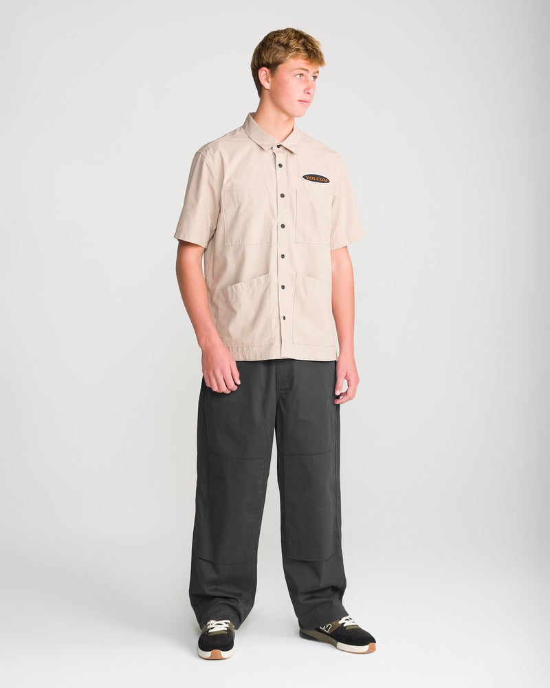 A young man stands against a plain background, wearing a beige short-sleeve button-up shirt with two front pockets and Volcom Men’s Stoneworks Utility Work Pants, which feature reinforced front-leg patches. He is looking slightly to the side with a neutral expression.