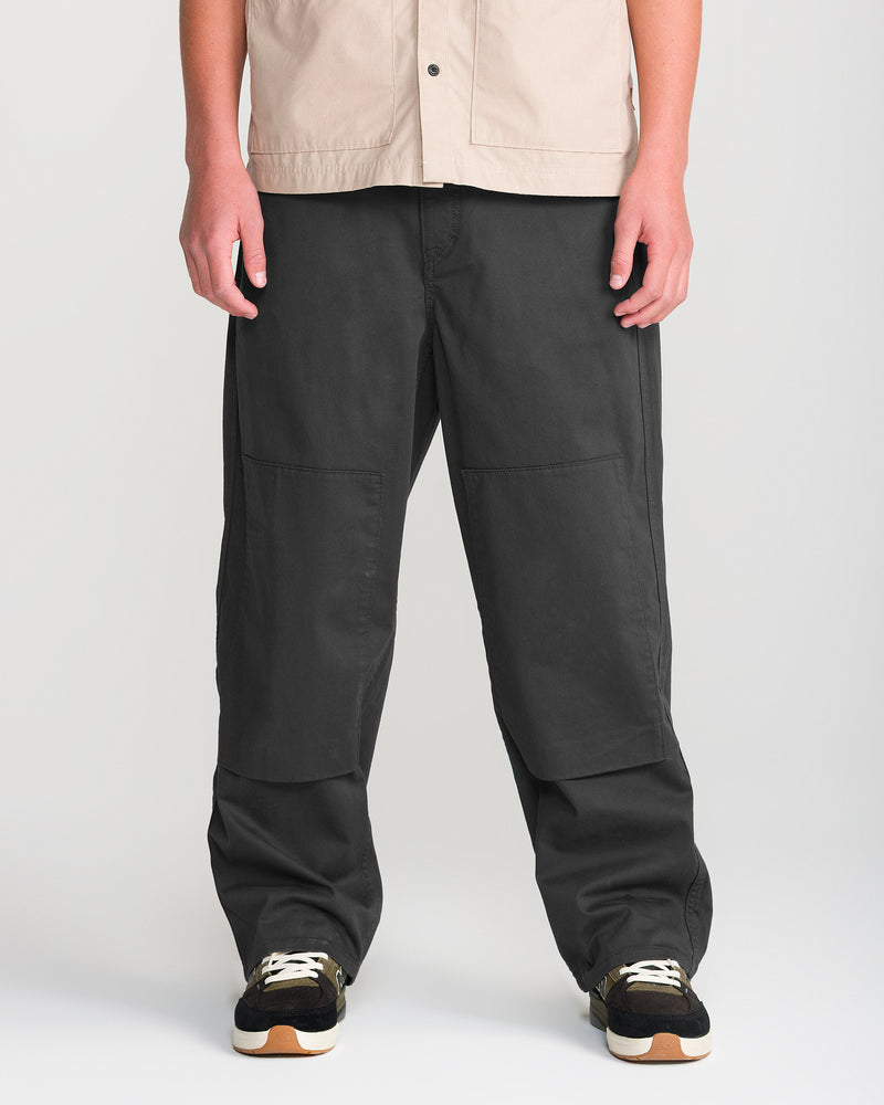 A person wearing a beige shirt, black sneakers with white laces, and Volcom Men’s Stoneworks Utility Work Pant made from stretch broken twill stands against a plain background. The dark gray Volcom pants feature reinforced front-leg patches for added durability.