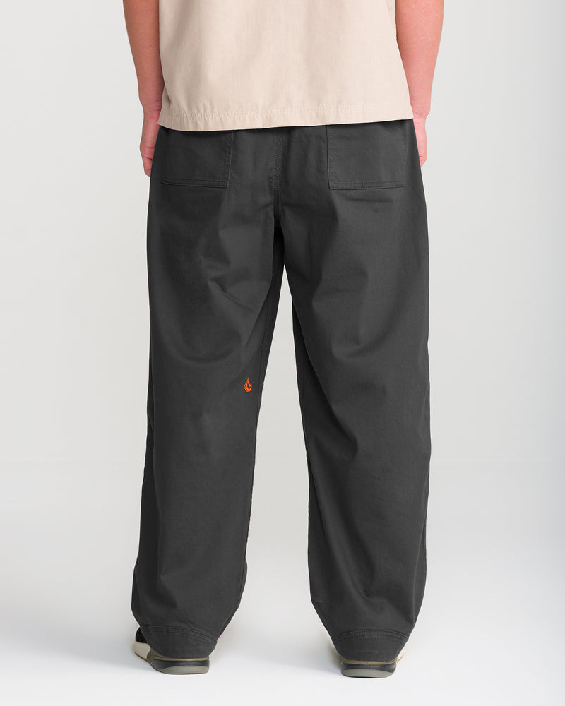 A person stands with their back to the camera, wearing Volcom’s Men’s Stoneworks Utility Work Pant in black—crafted from stretch broken twill fabric, featuring two back pockets and a small orange flame logo on the left leg—paired with a beige shirt and black shoes against a plain white background.