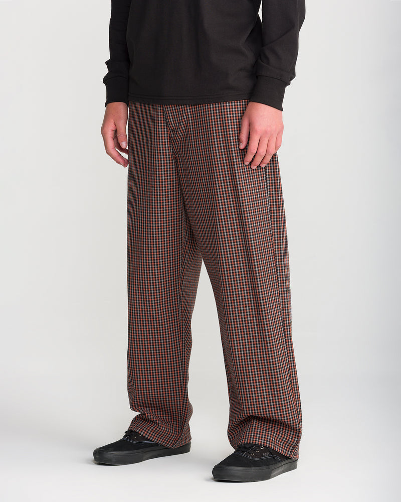 A person wearing Volcom Men’s Billow Plaid Pant, featuring low-rise, wide-leg trousers in an orange, black, and white box plaid twill, paired with black sneakers.