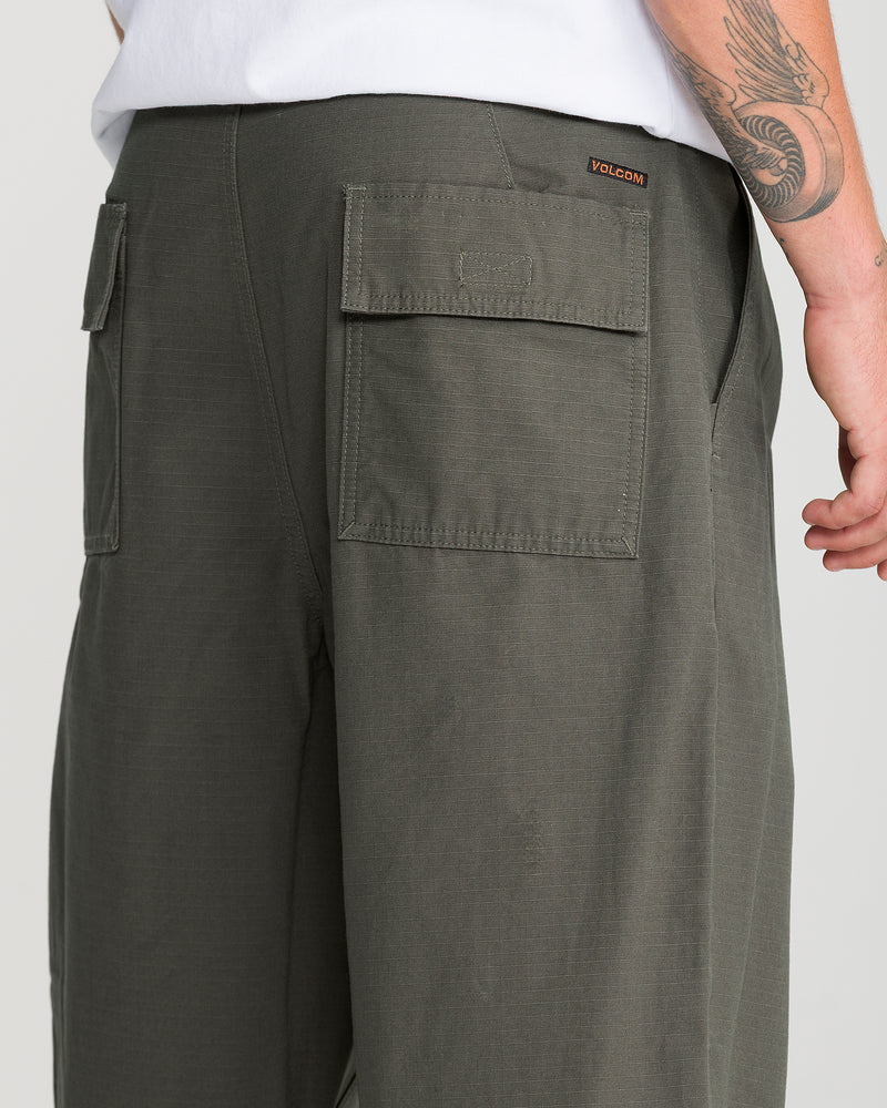 A person wears Volcom Men’s Skuds Pant in olive green cotton ripstop with large back pockets and a white shirt. Their tattooed right arm is partly visible, and the Volcom tag sits above the right pocket.