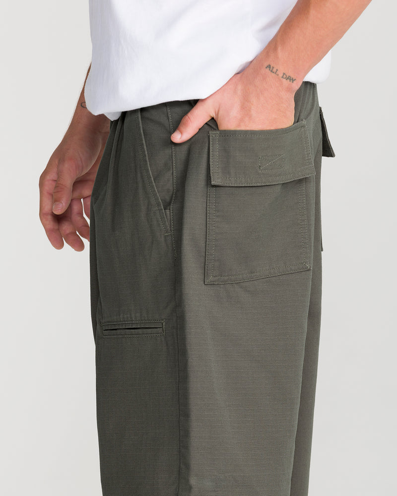 Wearing the Volcom Men’s Skuds Pant in olive green, a person stands with one hand in the cargo pocket. ALL DAY is tattooed on their wrist above a white shirt.