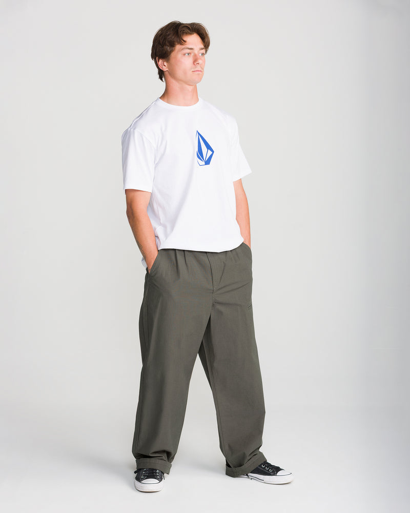A person stands with hands in pockets, wearing a white T-shirt, black sneakers, and Volcom Men’s Skuds Pant in olive-green cotton ripstop loose fit, against a plain light background.