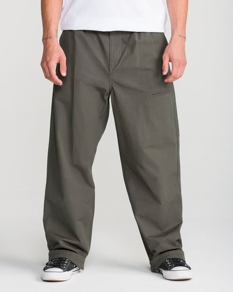 A person wears Volcom Men’s Skuds Pant in dark olive, paired with a white shirt and black-and-white sneakers, standing with arms relaxed against a plain light background. Only the lower body is visible.