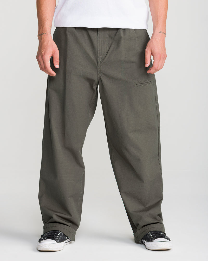 A person wears Volcom Men’s Skuds Pant in dark olive, paired with a white shirt and black-and-white sneakers, standing with arms relaxed against a plain light background. Only the lower body is visible.