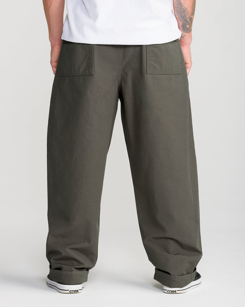 A person wears the Men’s Skuds Pant by Volcom in dark green, cuffed at the ankles, paired with a white shirt and white sneakers, facing away from the camera.