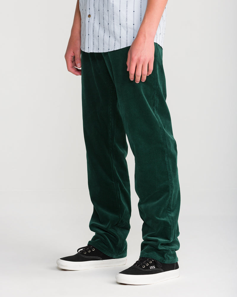 A person wearing Volcom Men’s Kinkade Cord Pant in dark green, a white and blue striped shirt, and black sneakers stands against a plain light background. Only the lower half is visible.