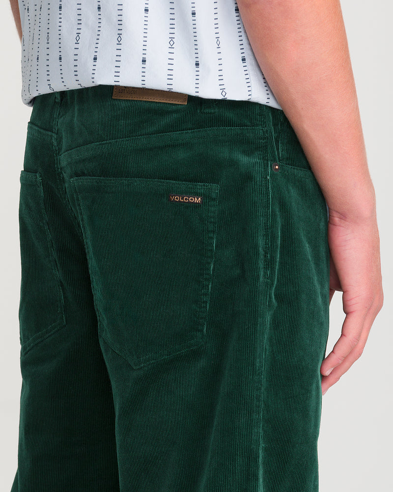A person wears green Men’s Kinkade Cord Pant by Volcom with straight legs, visible back pockets, and the Volcom logo on the right pocket, paired with a white shirt with thin blue stripes.