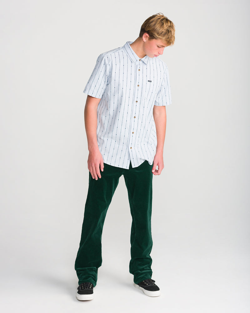 A person stands looking down, wearing a light blue striped short-sleeve shirt, Volcom Men’s Kinkade Cord Pant in dark green straight leg style, and black sneakers on a plain white background.