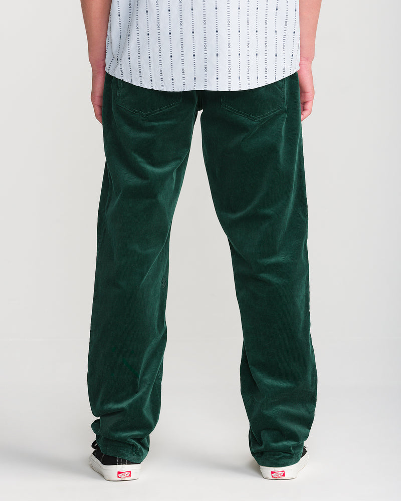 A person stands with their back to the camera, wearing a white patterned shirt, Volcom Men’s Kinkade Cord Pant in dark green, and white sneakers with red labels against a plain light background.