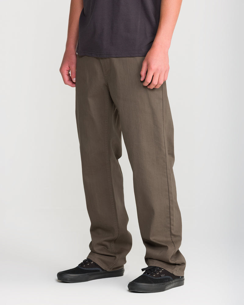 A person wearing Volcom Men’s Kinkade Slub Pant in brown, paired with a dark t-shirt and black sneakers, stands against a plain white background. Only their lower half is visible.