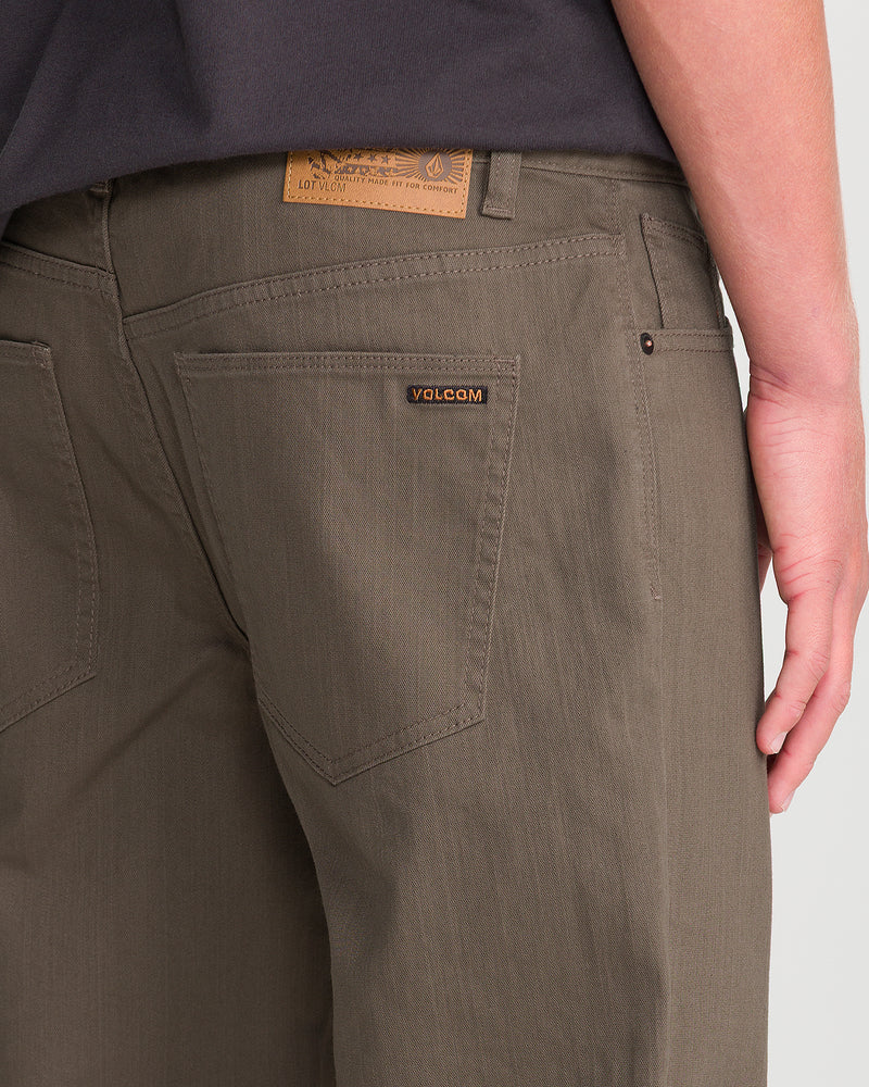 A person wears olive green Men’s Kinkade Slub Pant by Volcom, shown from the back with only the lower torso, waist, and right hand visible. The Volcom logo label is on the right back pocket of these straight leg pants.