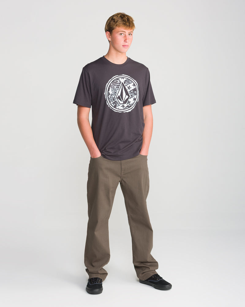 A young man stands against a white background, wearing a dark graphic Volcom T-shirt, Men’s Kinkade Slub Pant in khaki by Volcom, and black shoes, with his hands in his pockets.