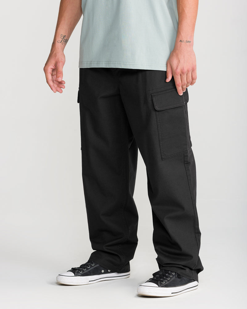 A person is shown from the lower body and arms, standing against a plain light background, wearing Volcom Men’s Billow Tapered Elastic Waist Cargo pants in black, styled with a light blue shirt and black low-top sneakers.