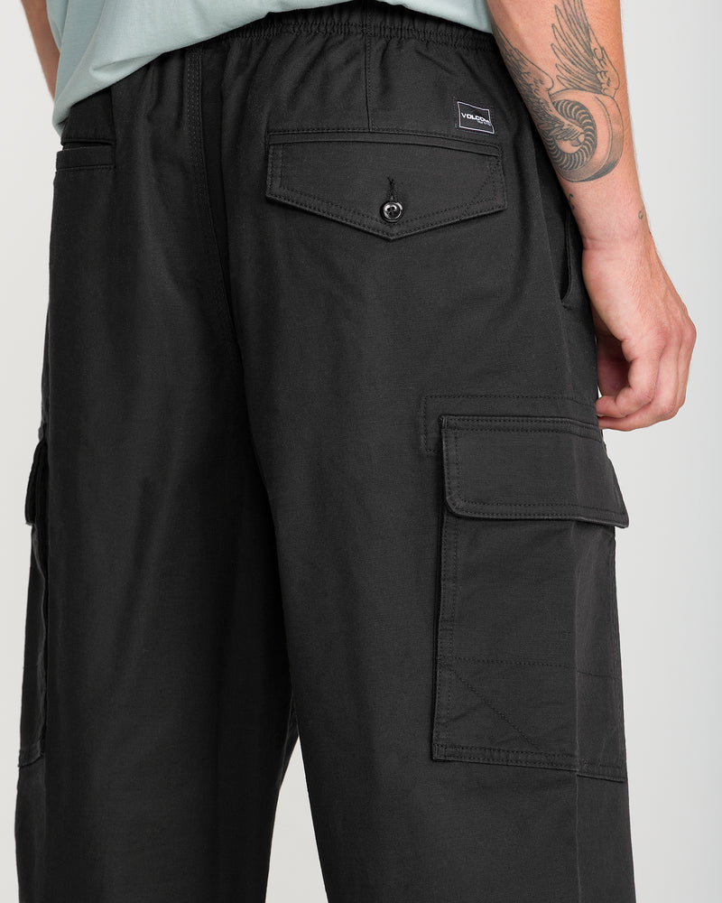 A person wearing Volcom Men’s Billow Tapered Elastic Waist Cargo pants in black, featuring flap pockets and a relaxed fit, paired with a light green shirt and showcasing an arm tattoo with a winged design. The image highlights the tapered leg of the pants from waist to knee.