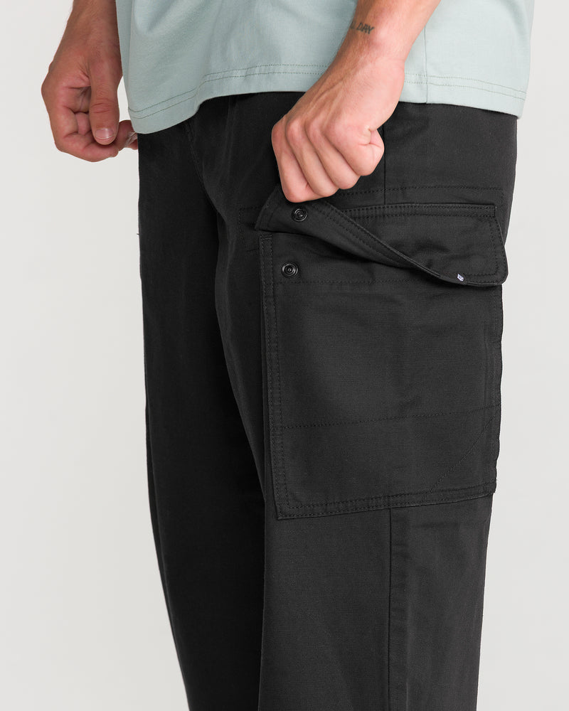 A person, their torso and hand visible, opens a buttoned side pocket on the Volcom Men’s Billow Tapered Elastic Waist Cargo pants in black; a light green shirt is partially shown.