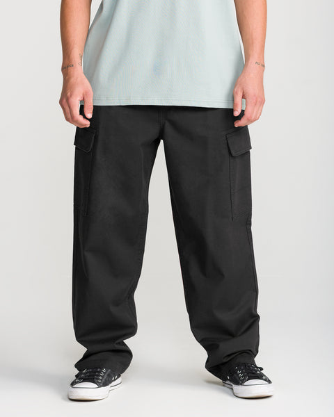 A person is shown from the waist down, wearing Volcom Men’s Billow Tapered Elastic Waist Cargo pants in black, paired with a light green T-shirt and black sneakers, standing against a plain light background.