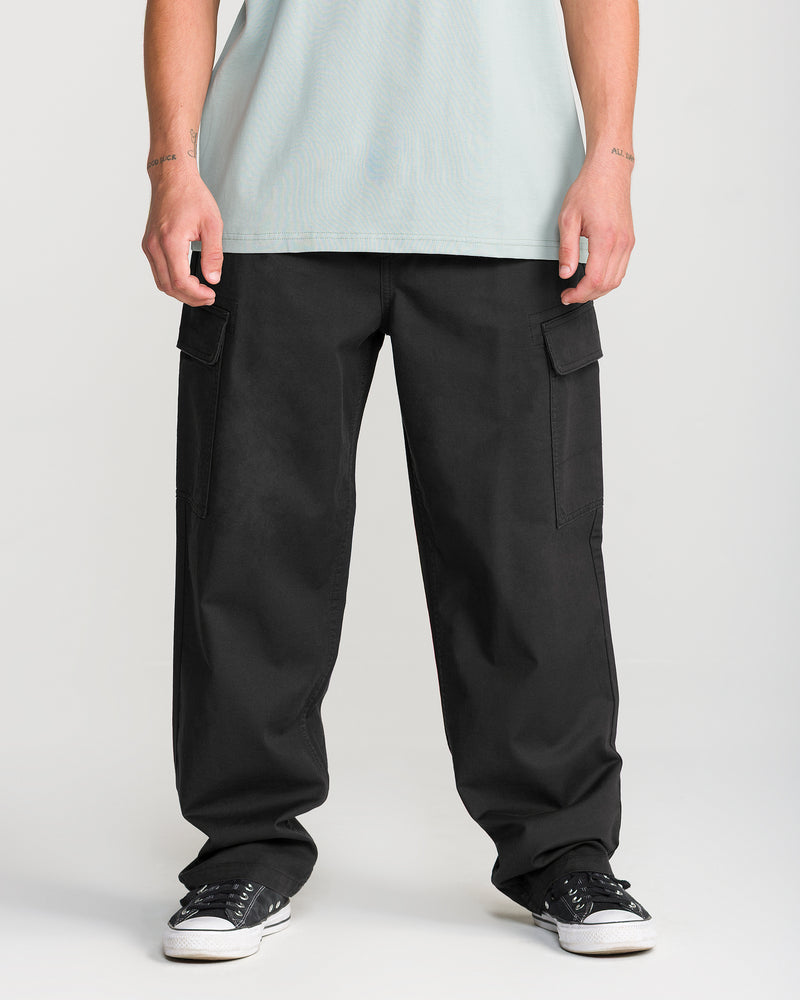 A person is shown from the waist down, wearing Volcom Men’s Billow Tapered Elastic Waist Cargo pants in black, paired with a light green T-shirt and black sneakers, standing against a plain light background.