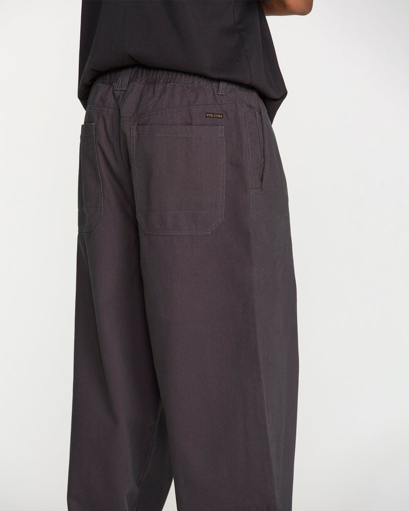 A person is seen from behind wearing Volcom Men's Chillow Pleat Elastic Waist Pants in a loose charcoal gray cotton twill, featuring visible rear pockets, paired with a black shirt; the image focuses on the lower back and upper legs against a plain background.