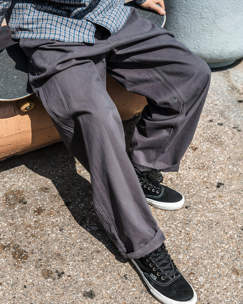 A person wearing Volcom Men’s Chillow Pleat Elastic Waist Pants in soft gray, paired with black sneakers and a plaid shirt, sits on a bench outdoors. The image is cropped to show only the lower half of their body.