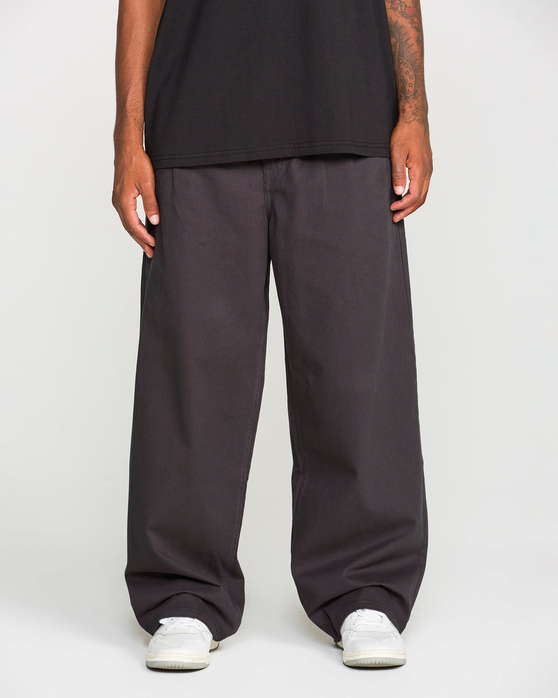 A person, shown from the waist down against a plain white background, is wearing Volcom Men's Chillow Pleat Elastic Waist Pants in a loose dark gray fit, paired with a black shirt and white sneakers.
