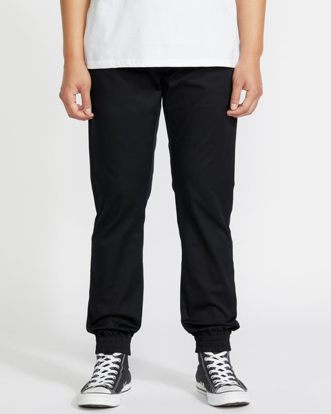 A person, visible only from the waist down, wears Volcom Men's Frickin Slim Joggers in black—featuring elastic cuffs and made with REPREVE recycled fibers—paired with a white shirt and black-and-white Converse sneakers against a white background.
